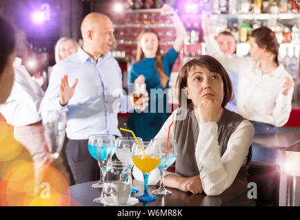 Sad woman sitting alone at office party in nightclub in background with cheerful workmates Stock Photo