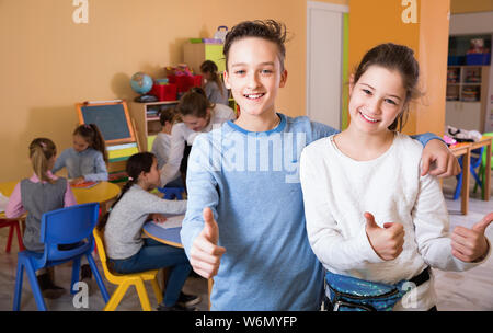 Two satisfied  positive   children standing in schoolroom on background with pupils studying with teacher Stock Photo