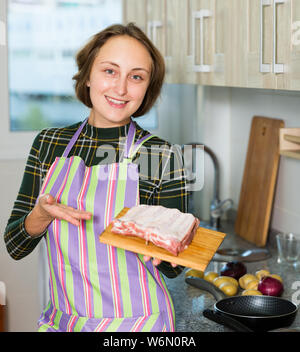 Smiling young woman ready to cook pork ribs, standing near kitchen table Stock Photo