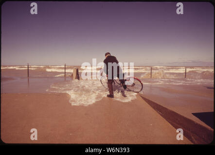 WAVE CRASHES OVER SEAWALL, TAKING AN ELDERLY BICYCLE RIDER BY SURPRISE Stock Photo