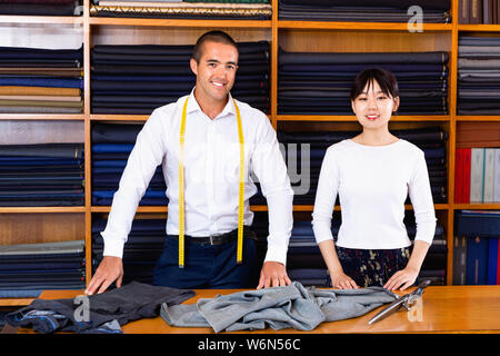 Portrait of confident cheerful positive smiling male tailor with young female assistant standing next to cutting table in atelier Stock Photo