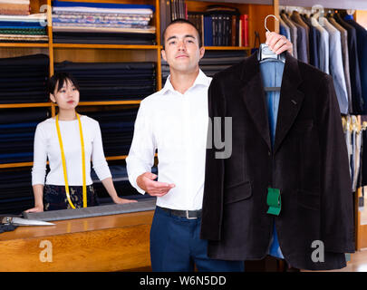 Satisfied male client of sewing workshop showing his new suit jacket on background of young Chinese female tailor Stock Photo