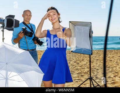 Young cheerful positive attractive female model delighted with professional photo shooting on seaside with male photographer Stock Photo