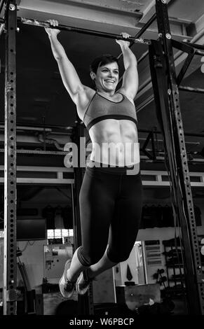 A young beautiful woman with abs is doing toes to bar at the horizontal bar. The short haired sporty athlete is doing a functional fitness workout. Stock Photo