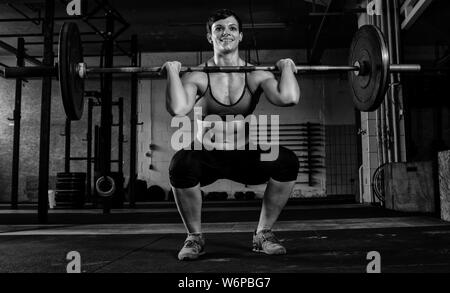 A fit short haired woman with Sixpack is doing weight lifting with a barbell in a gym. The muscular woman is doing front squats. Stock Photo