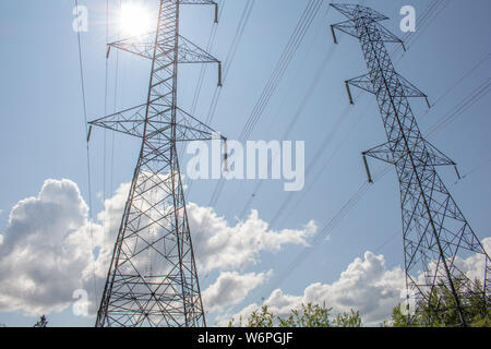 A pair of electrical transmission towers suspend cables and stand tall against a blue sky, with the sun shining through from behind. Stock Photo