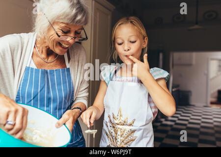 Senior woman in apron making batter for cake. Little girl tasting cake batter standing in kitchen with grandmother. Stock Photo