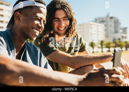 Young man with his friend looking at a mobile phone and smiling outdoors. Two young men sitting outdoors using a smart phone. Stock Photo