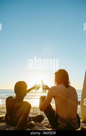 Rear view of two young men relaxing on the beach with beers. Young men toasting beers on the beach during sunset. Stock Photo
