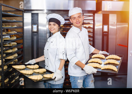 baker boy and girl with baking sheets with raw dough on the background of an industrial oven in a bakery. Stock Photo