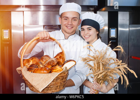 bakers boy and girl with a baking basket and spikelets of wheat against the background of an industrial oven in a bakery. Stock Photo