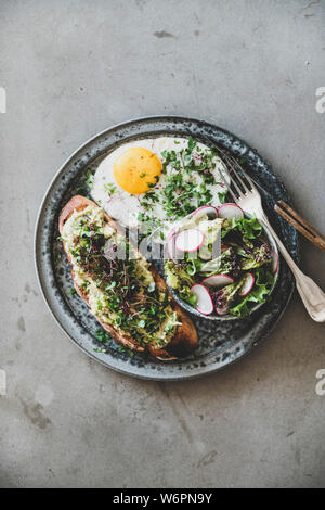 Healthy trendy breakfast. Flat-lay of avocado toast on sourdough bread with herbs and sprouts, fried egg with coriander and salad in plate over grey c Stock Photo