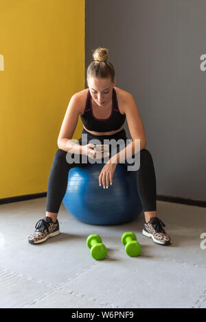 Sport woman sitting, resting and watching her mobile after workout or exercise in fitness gym. Stock Photo