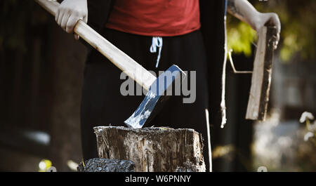 The woodcutter guy , dressed in black clothes and a red t-shirt, holds a sharp long axe in one hand, sticking it into a stump, and in the other hand h Stock Photo