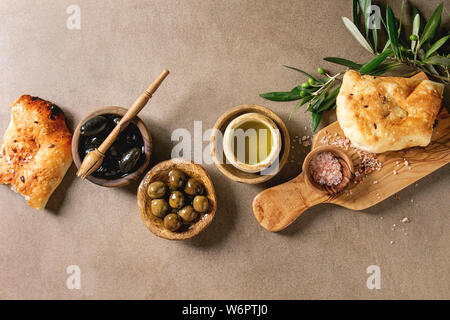Variety of green and black whole olives in olive oil served in bowls with fresh baked ciabatta bread, pink salt and young olive wood branches over bro Stock Photo