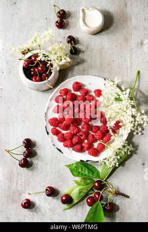 Fresh cherry and raspberry berries in ceramic mug and plate, elderflowers, jug of cream over gray texture background. Flat lay, space Stock Photo