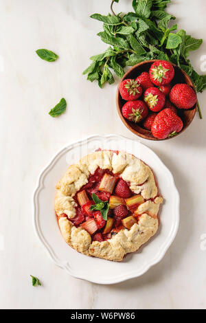 Home baking summer berry biscuit pie with strawberry and rhubarb, served in plate with fresh strawberries and mint over white marble background. Flat Stock Photo