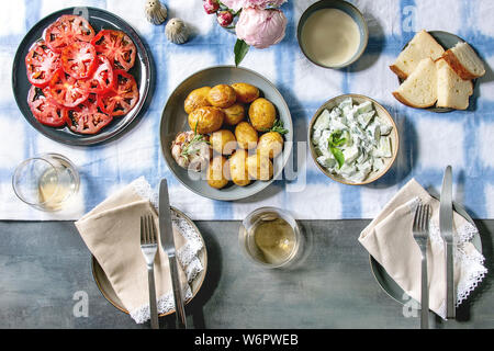 Summer vegetarian dinner table for family or friends. Young baked potatoes, tomato carpaccio, cucumber salad, bread, wine, sauce and flowers over line Stock Photo