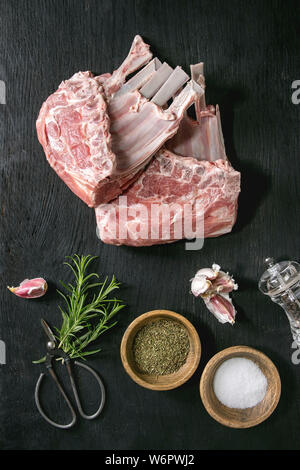 Raw uncooked rack of lamb with salt, herbs rosemary, pepper and garlic on black burned wooden background. Flat lay Stock Photo