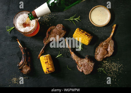 Grilled bbq rack of lamb with sweet corn cobs, rosemary and cheese sauce, glass of lager beer, flowing from bottle, over black burned wooden backgroun Stock Photo