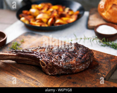 Grilled angus beef tomahawk steak on a cutting board with roasted potatoes in wrought iron pan Stock Photo