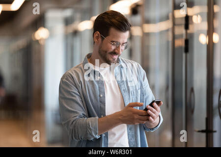 Happy young businessman using smart phone standing in office hallway Stock Photo