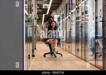 Excited diverse colleagues laughing riding on chair in office hallway Stock Photo