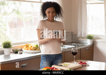 African woman standing in kitchen distracted from cooking using cellphone Stock Photo