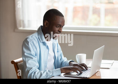 African millennial guy sitting at table typing on laptop Stock Photo