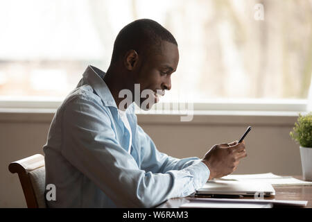 Side view african guy sit at table smiling using smartphone Stock Photo