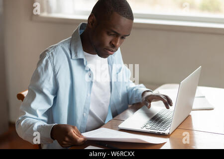 African man sitting at table paying household bills using laptop Stock Photo