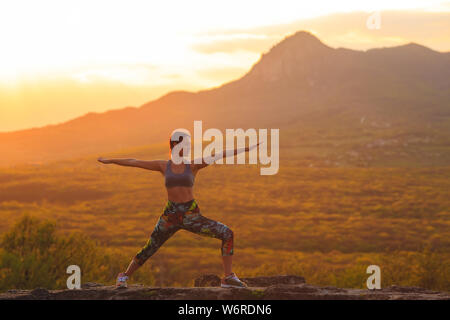 Silhouette of young woman practicing yoga or pilates at sunset or sunrise in beautiful mountain location, doing lunge exercise, standing in Warrior Stock Photo
