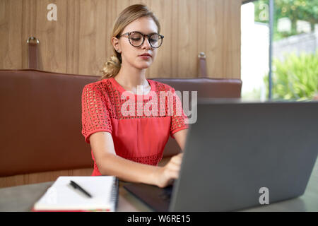 Closeup lifestyle portrait of professional blonde caucasian millennial student wearing glasses taking notes on laptop computer in a modern restaurant Stock Photo