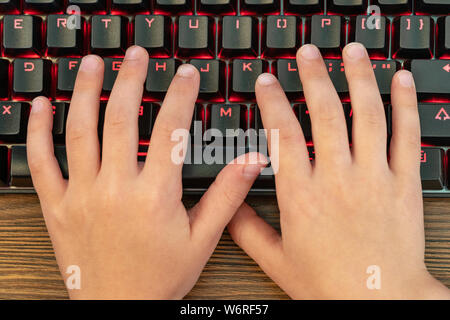 two children hands are typing on a black keyboard with red backlight lying on a wooden table Stock Photo