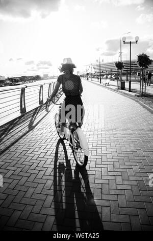 young curly hair woman in hat and dress moves on bike near river at city, rear view, sunset time, monochrome Stock Photo