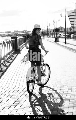 young curly hair woman in hat and dress moves on bike near river at city, rear view, sunset time, looking back, monochrome Stock Photo
