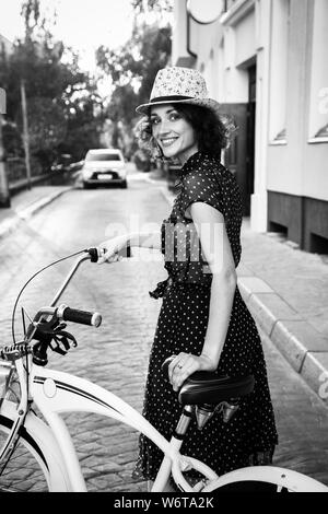 young curly hair woman in hat and dress posing with bike at old european city, sunset time, looking at camera, smiling, monochrome Stock Photo