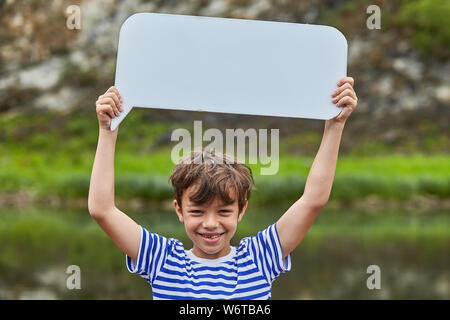 Little white boy is holding blank steep of paper and smiling, he is in forest and there is a river behind him, ecotourism. Stock Photo