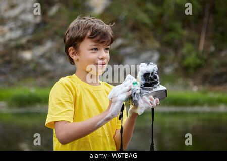 White boy is washing  digital camera, which is covered by foam, the boy is standing near river in forest or park and smiling, eco tourism. Stock Photo