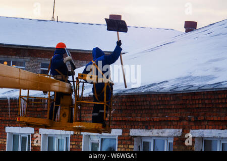 Workers in overalls and orange helmets on the crane basket remove icicles from roof of the house on a winter day - cleaning the roofing, utility servi Stock Photo