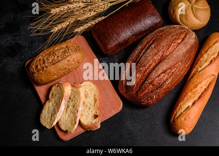 Fresh fragrant bread with grains and cones of wheat against a dark background. Assortment of baked bread on wooden table background Stock Photo