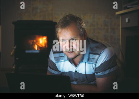 man watching content on a laptop in the evening at home by the fireplace Stock Photo