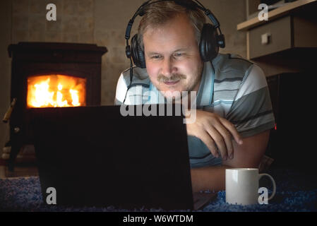 A man with headphones uses a laptop at night on the floor of the room by the fireplace Stock Photo