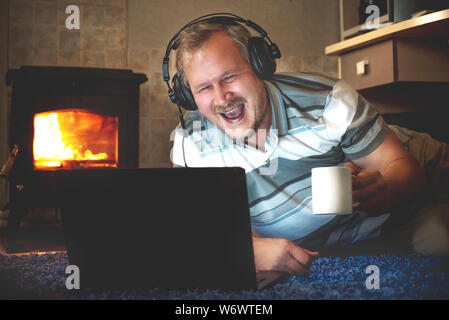 Cheerful man watching online content on a laptop at night on the floor of the room by the fireplace Stock Photo