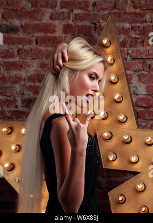 Beautiful emotional fun blond cheeky teen girl showing the tongue and rock cool sign the two fingers on bricks wall and yellow star background. Closeu Stock Photo