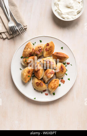 Fried dumplings stuffed with potato and meat (pierogi) sprinkled with bacon and  parsley and sour cream on white wooden table, copy space. Stock Photo