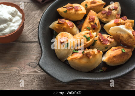 Fried dumplings stuffed with potato and meat (pierogi) sprinkled with bacon and  parsley and sour cream on wooden table, close up. Stock Photo
