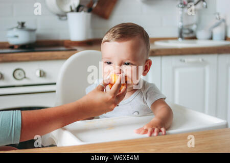 Charming little baby boy eating first food peach at the kitchen. Mom feeds child Stock Photo