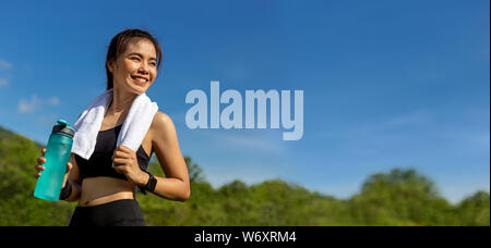 Happy beautiful young Asian woman with her white towel over her neck, standing smiling and holding her water bottle to drink after her morning exercis Stock Photo