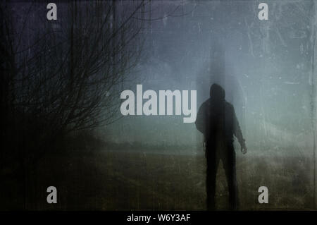 An eerie silhouette of a lone hooded figure in a field on a winters night. With a dark, spooky blurred abstract, grunge effect edit. Stock Photo